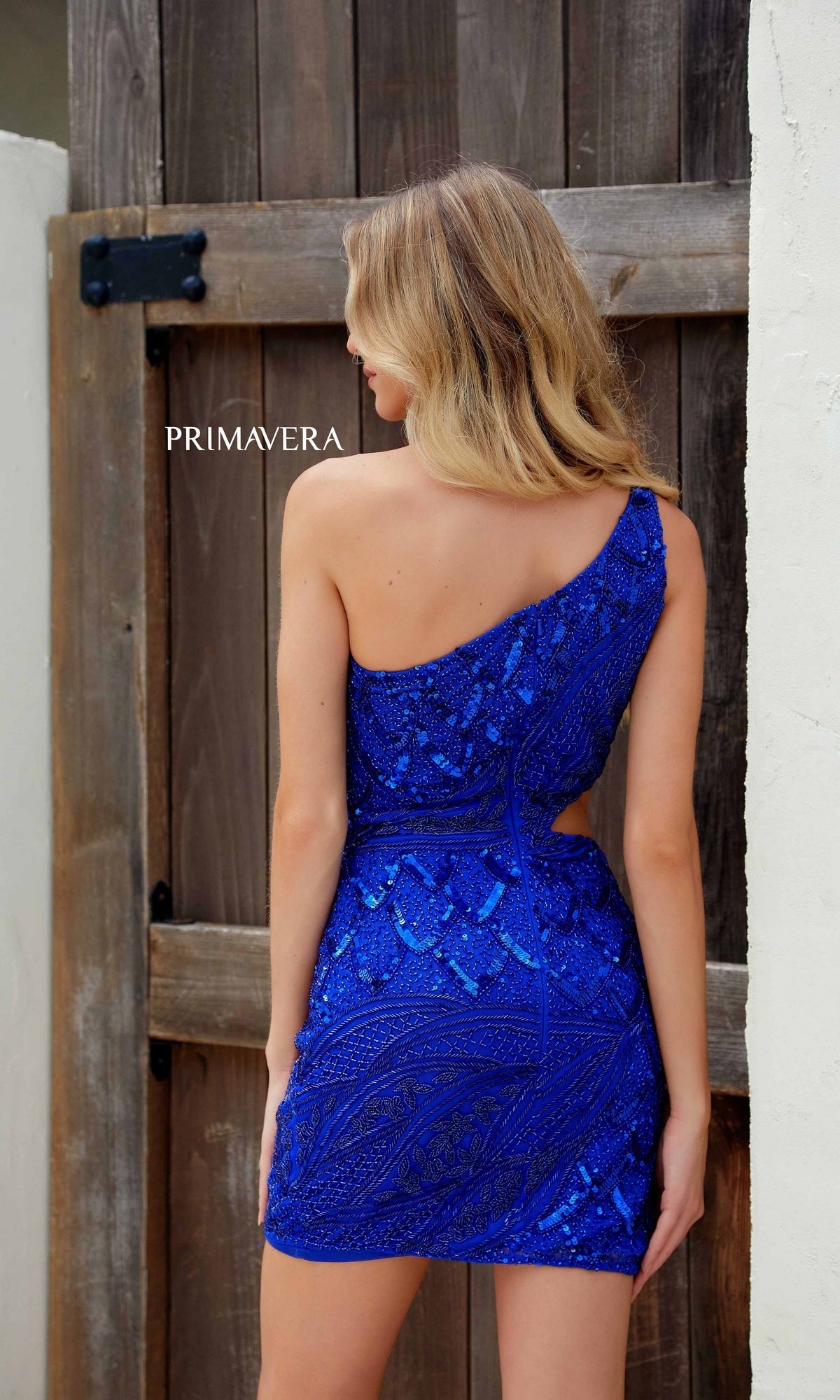 A woman stands with her back to the camera wearing the Primavera 4240 Cut-Out Short Beaded Hoco Dress, a sparkling blue one-shoulder mini perfect for any special occasion, in front of a wooden gate.